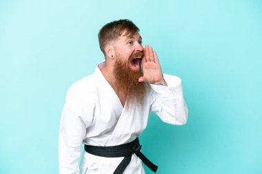 Uzun sakallı kızıl saçlı adam mavi arka planda karate yapıyor. Ağzı sonuna kadar açık bağırıyor.