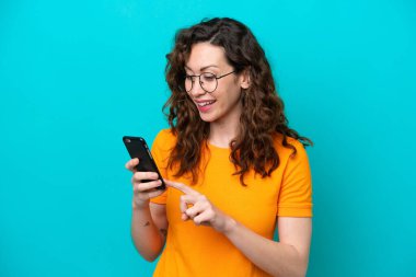 Mavi arka planda izole edilmiş genç beyaz kadın cep telefonuyla mesaj ya da e-posta gönderiyor.