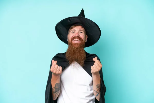 Young wizard in halloween isolated on blue background making money gesture