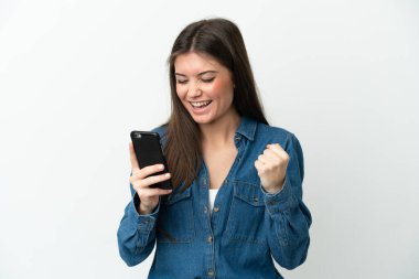 Beyaz arka planda izole edilmiş genç beyaz kadın cep telefonu kullanıyor ve zafer jesti yapıyor.