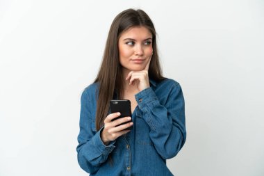 Beyaz arka planda cep telefonu kullanan ve düşünen genç beyaz bir kadın.