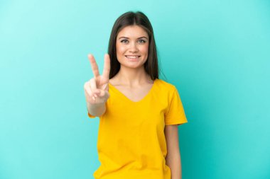 Mavi arka planda izole edilmiş genç beyaz kadın gülümsüyor ve zafer işareti gösteriyor.