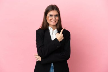 Pembe arka planda izole edilmiş genç beyaz kadın bir ürünü sunmak için kenarı işaret ediyor.