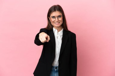 Genç beyaz kadın pembe arka planda izole edilmiş mutlu bir ifadeyle ön tarafı işaret ediyor.