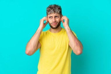 Mavi arka planda izole edilmiş yakışıklı beyaz adam hayal kırıklığına uğramış ve kulaklarını kapatmış.