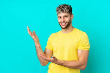 Mavi arka planda izole edilmiş yakışıklı beyaz adam, davet edilmek için ellerini yana uzatıyor.