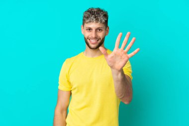 Mavi arka planda izole edilmiş yakışıklı beyaz adam parmaklarıyla beşi sayıyor.