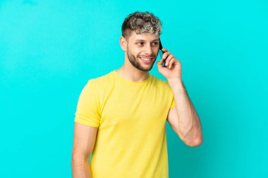 Mavi arka planda izole edilmiş yakışıklı beyaz bir adam cep telefonuyla sohbet ediyor.