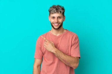 Mavi arka planda izole edilmiş yakışıklı beyaz adam bir ürünü sunmak için kenarı işaret ediyor.