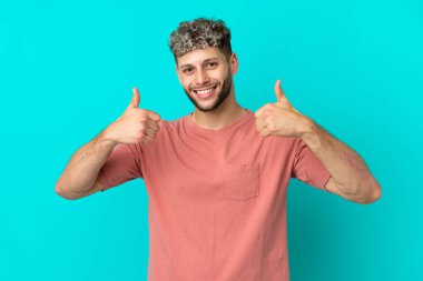 Genç, beyaz, mavi arka planda izole edilmiş genç bir adam baş parmak işareti yapıyor.
