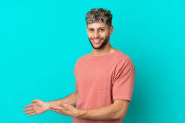 Mavi arka planda izole edilmiş yakışıklı beyaz adam, davet edilmek için ellerini yana uzatıyor.