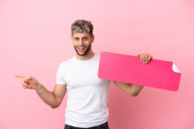 Pembe arka planda izole edilmiş yakışıklı beyaz bir adam boş bir levha tutuyor ve onu işaret ediyor.