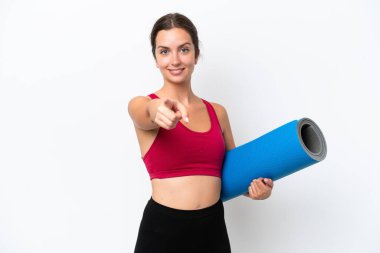 Beyaz arka planda izole edilmiş bir minderi tutarak yoga derslerine giden beyaz, neşeli bir ifadeyle öne bakan genç bir kadın.