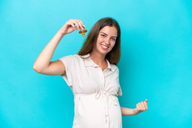 Genç beyaz kadın mavi arka planda izole edilmiş hamile ve emzik taşıyor ve zaferi kutluyor.