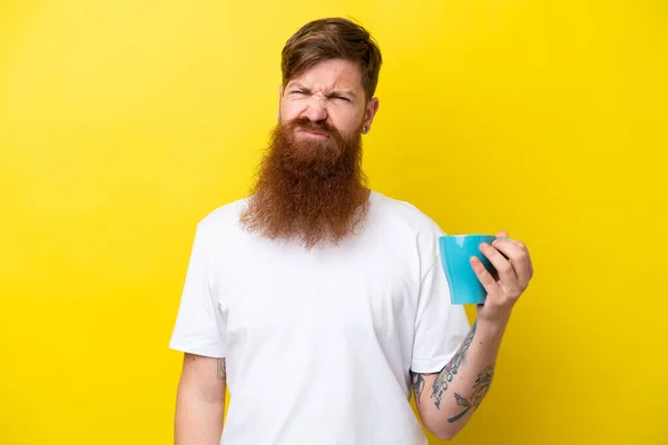 Redhead man with beard holding a mug isolated on yellow background with sad expression