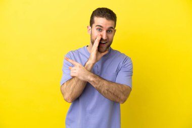 Sarışın yakışıklı, izole edilmiş sarı arka planda bir ürün sunması ve bir şeyler fısıldaması için bir tarafa bakıyor.