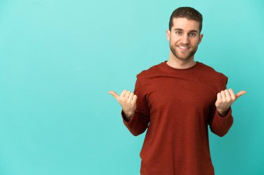 Yakışıklı sarışın, soyutlanmış mavi arka planda, başparmakları havada ve gülümsüyor.