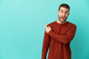 Yakışıklı sarışın, soyutlanmış mavi arka planda, şaşırmış ve işaret eden bir adam.