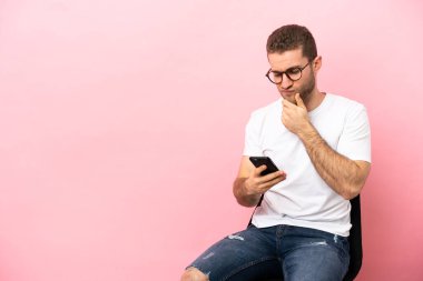 İzole edilmiş pembe arka planda bir sandalyede oturan genç adam düşünüyor ve bir mesaj gönderiyor.