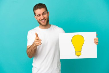Sarışın yakışıklı, izole edilmiş mavi arka planda elinde ampul simgesi olan ve ön tarafı gösteren bir levha tutan adam.