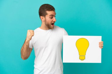 Sarışın, soyutlanmış, mavi arka planı olan yakışıklı bir adam. Elinde ampul simgesi olan bir pankart var ve zaferi kutluyor.