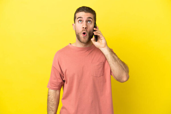 Handsome blonde man using mobile phone over isolated background looking up and with surprised expression