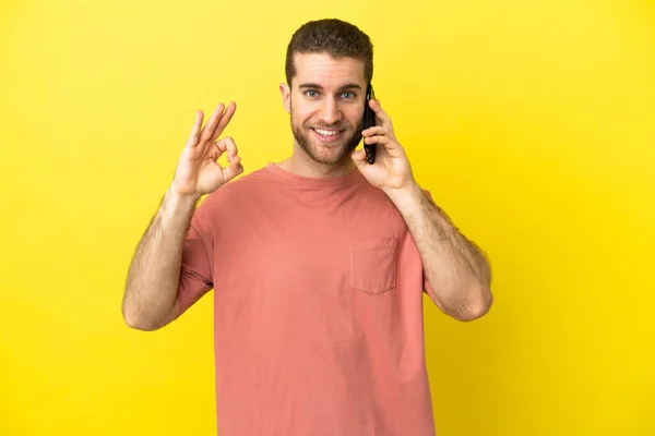 Przystojny Blondynka Człowiek Pomocą Telefonu Komórkowego Odosobnionym Tle Pokazuje Znak — Zdjęcie stockowe