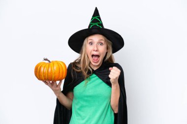 Cadı gibi giyinmiş genç sarışın bir kadın beyaz arka planda izole edilmiş bir balkabağı tutuyor. Kazanan pozisyonda zaferi kutluyor.