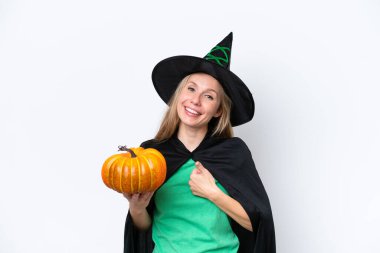 Beyaz arka planda izole edilmiş balkabağı tutan cadı gibi giyinmiş genç sarışın bir kadın baş parmak işareti yapıyor.