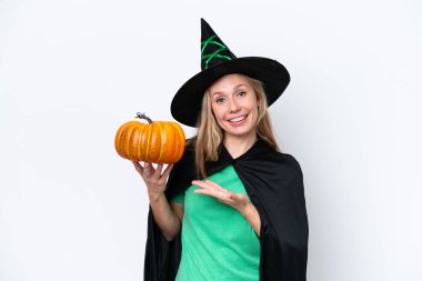 Cadı gibi giyinmiş, balkabağı tutan genç sarışın kadın beyaz arka planda izole edilmiş ve davet edilmek için ellerini yana uzatıyor.