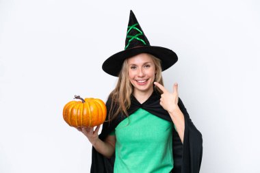 Beyaz arka planda izole edilmiş balkabağı tutan cadı gibi giyinmiş genç sarışın bir kadın baş parmak işareti yapıyor.