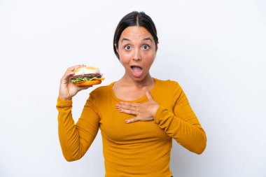 Beyaz arka planda yüzü sürpriz bir ifadeyle izole edilmiş bir burger tutan genç İspanyol kadın.