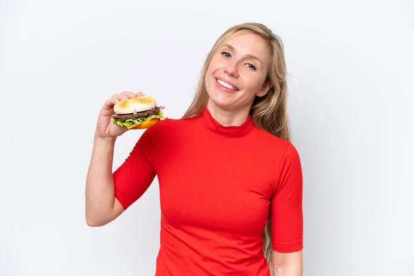 Młoda Blondynka Trzyma Hamburgera Odizolowany Białym Tle Uśmiechając Się Dużo — Zdjęcie stockowe