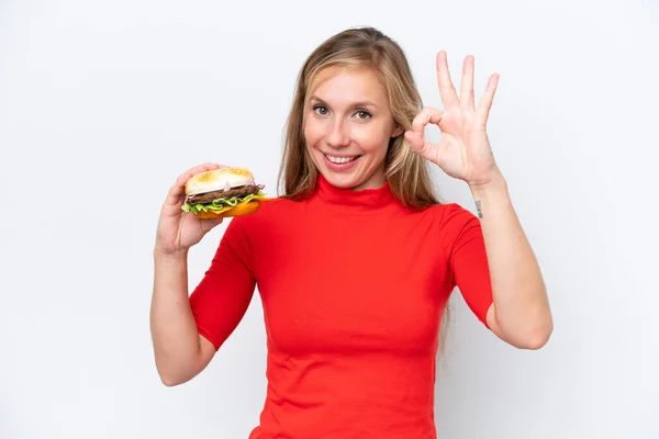 Młoda Blondynka Trzyma Hamburgera Białym Tle Pokazuje Znak Palcami — Zdjęcie stockowe