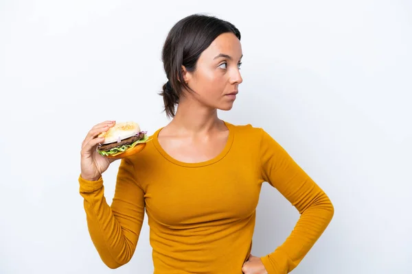 Młoda Latynoska Trzyma Hamburgera Białym Tle Patrząc Boku — Zdjęcie stockowe