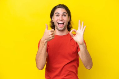 Beyaz tenli yakışıklı adam sarı arka planda izole edilmiş tamam işareti ve baş parmak hareketi gösteriyor.