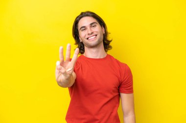 Sarı arka planda izole edilmiş beyaz yakışıklı adam mutlu ve parmaklarıyla üçe kadar sayıyor.