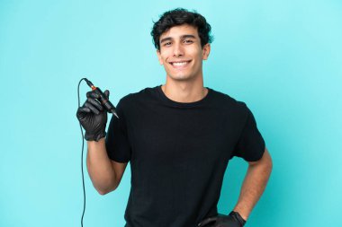 Dövmeci Arjantinli adam mavi arka planda izole edilmiş, kolları kalçasında ve gülümsüyor.