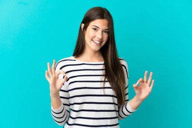 İzole edilmiş mavi arka plan üzerinde Brezilyalı genç bir kız iki eliyle tamam işareti gösteriyor.