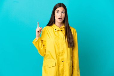 İzole edilmiş mavi arka planda, yağmura dayanıklı bir ceket giyen Brezilyalı genç kız parmağını kaldıracak bir fikir düşünüyor.