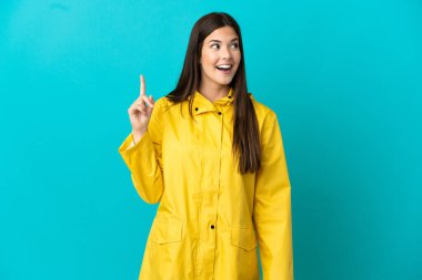 İzole edilmiş mavi arka planda yağmura dayanıklı bir ceket giyen Brezilyalı genç kız çözümü fark etmek için parmağını kaldırıyor.