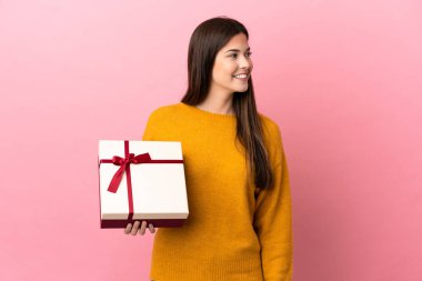 İzole edilmiş pembe arka planda elinde bir hediye tutan Brezilyalı genç kız yana bakıyor ve gülümsüyor.