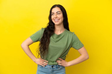 Sarı arka planda izole edilmiş genç beyaz kadın kolları kalçasında ve gülümsüyor.