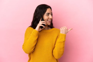 Young caucasian woman using mobile phone isolated on pink background pointing to the side to present a product