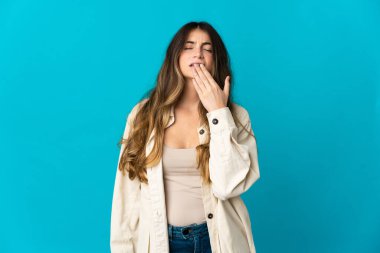 Genç beyaz kadın mavi arka planda izole edilmiş esniyor ve geniş bir ağzı eliyle kapatıyor.