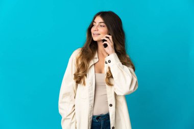 Mavi arka planda izole edilmiş genç beyaz bir kadın cep telefonuyla sohbet ediyor.