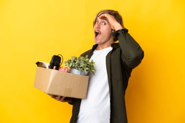 Sarışın adam, sarı arka planda izole edilmiş eşyalarla dolu bir kutuyu kaldırırken bir yandan yan tarafa bakarken sürpriz jest yapıyor.