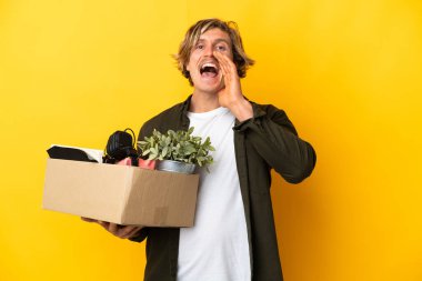 Sarışın adam, sarı arka planda izole edilmiş şeylerle dolu bir kutuyu alıp ağzı açık bağırarak hareket ediyor.
