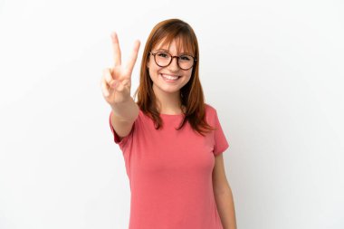 Kızıl saçlı kız beyaz arka planda izole edilmiş gülümsüyor ve zafer işareti gösteriyor.