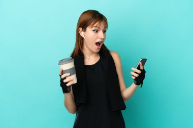 Redhead sport girl isolated on blue background holding coffee to take away and a mobile
